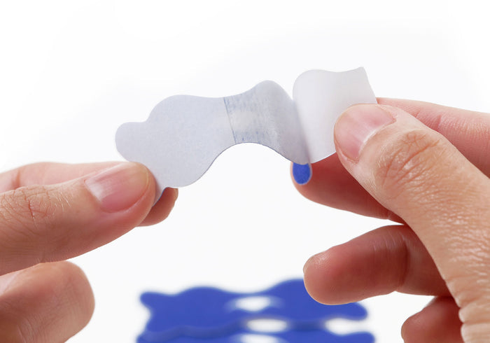 Close-up shot of hands peeling a white protective backing paper off a blue Manta nasal strip.