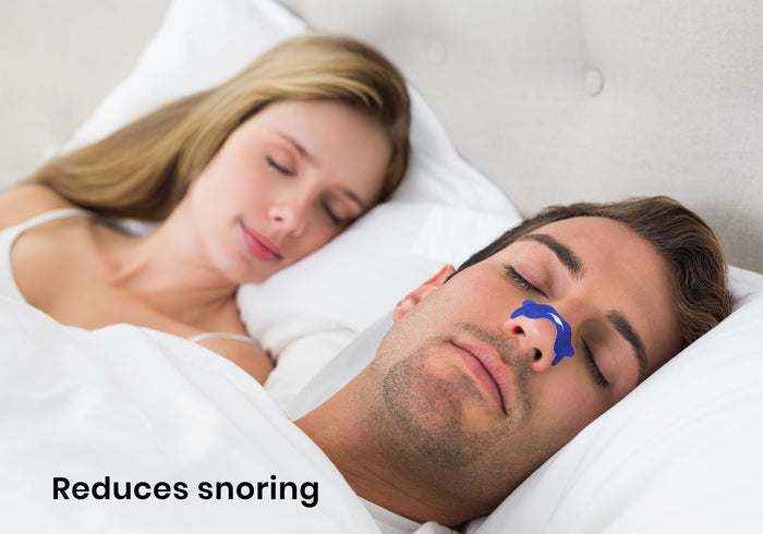 A man sleeping in bed with a blue nasal strip on his nose next to a sleeping woman. Text overlay: "Reduces snoring".